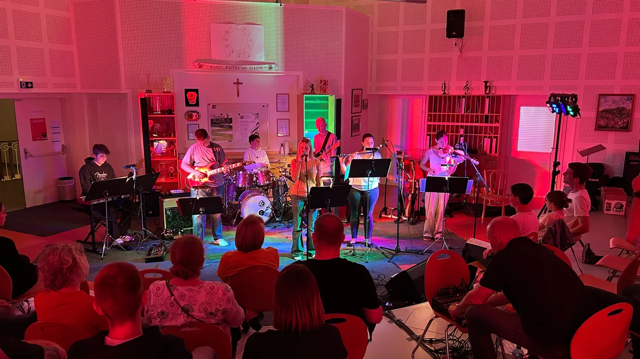 Eine Band spielt auf der Bühne mit einer roten und grünen Lichtshow. Das Publikum sitzt auf roten Stühlen und beobachtet die Band. Ein Kreuz hängt an der Wand über der Bühne.