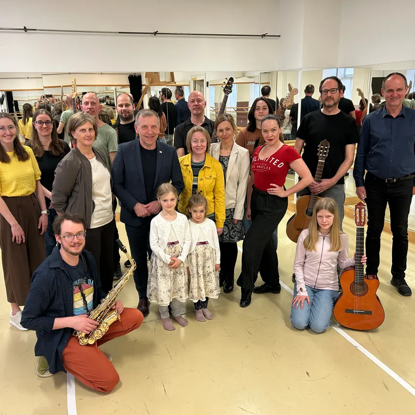 Eine Gruppe von Menschen posiert für ein Foto in einem Raum mit Gitarren. Einige halten Gitarren, andere posieren mit Instrumenten. Ein Mann spielt eine Saxophon und kniet.