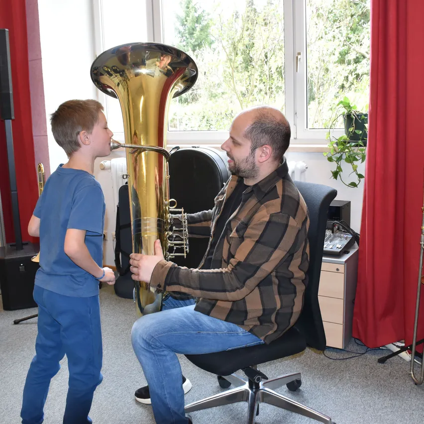 Ein Mann in einem karierten Hemd und Jeans lehrt einen jungen Jungen, wie man in einem Musikzimmer Tuba spielt.