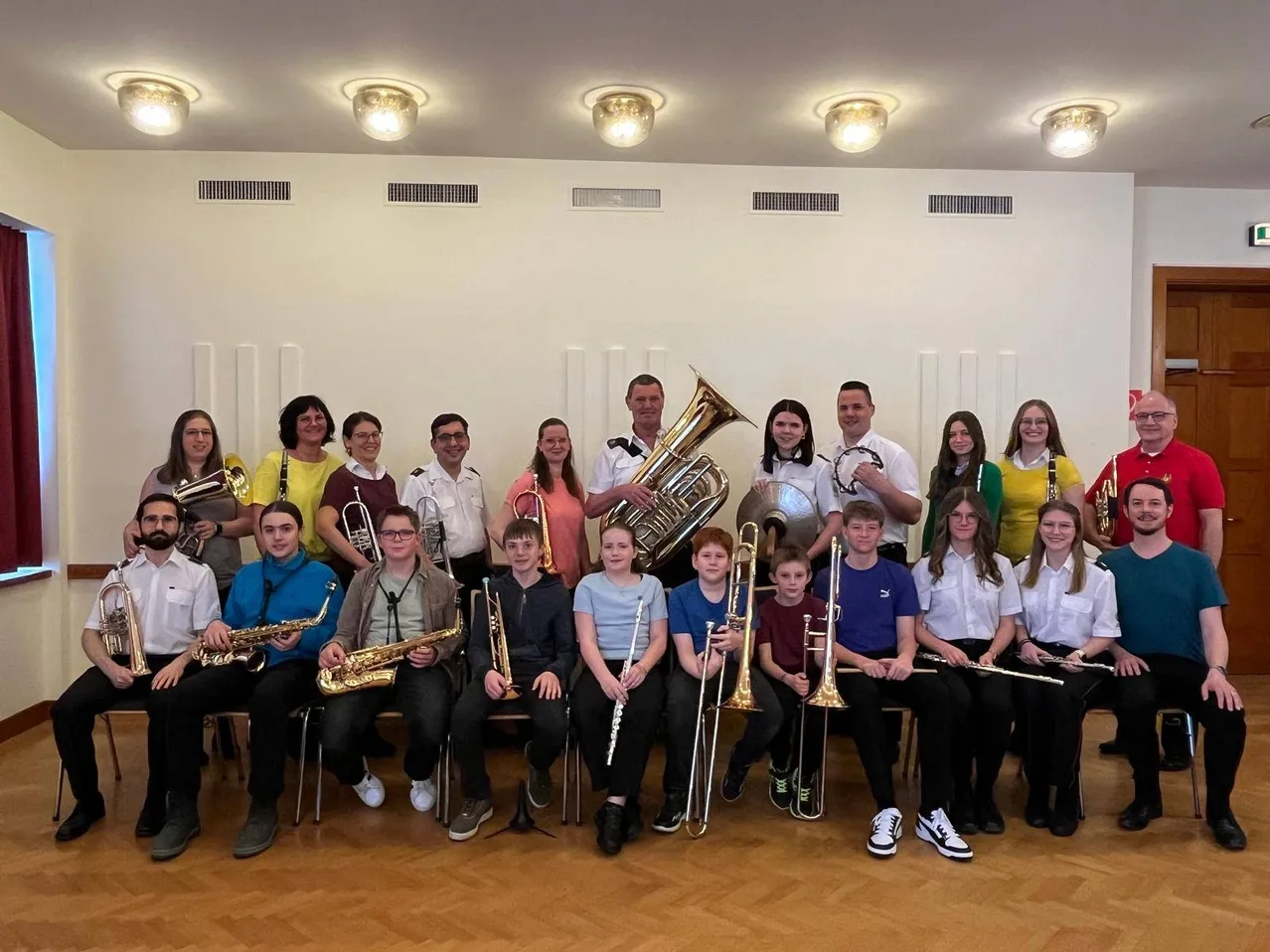 Eine Gruppe junger Musiker posiert mit ihren Instrumenten in einem gut beleuchteten Raum. Die Instrumente umfassen Trompeten, Saxophone, Posaunen und Flöten.