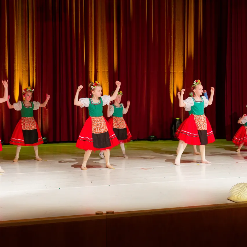 Eine Gruppe junger Mädchen in traditionellen Kostümen tanzt auf einer Bühne mit einem roten Vorhang im Hintergrund.