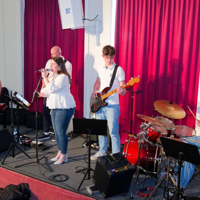 Eine Band tritt auf einer Bühne mit roten Vorhängen auf. Die Frau singt in ein Mikrofon, während der Mann Gitarre spielt. Rechts sind Trommeln und Becken.