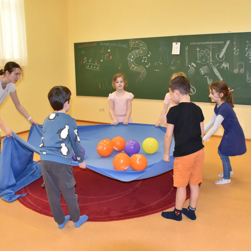 Mehrere Kinder spielen mit einem blauen Tuch voller Ballons, beaufsichtigt von einem Erwachsenen in einem Klassenzimmer mit einer musikalischen Instrumentenzeichnung auf der Tafel.