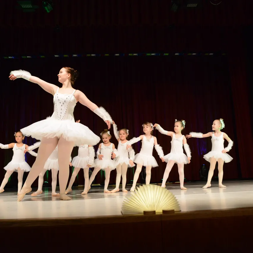 Eine Ballerina führt eine Gruppe junger Mädchen in einer Ballettaufführung auf der Bühne an, mit roten Vorhängen im Hintergrund.