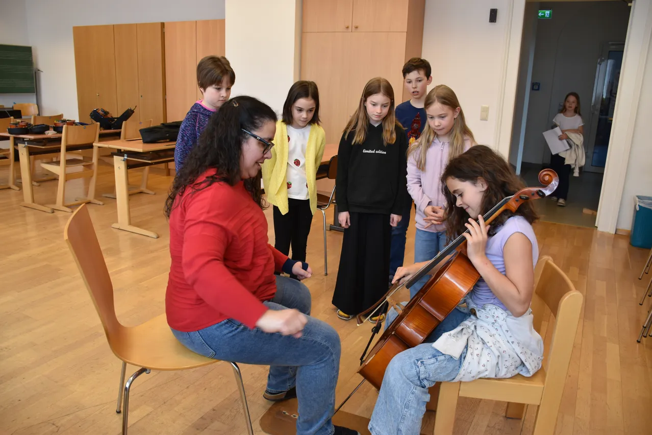 Eine Frau in einem roten Shirt und mit Brille lehrt ein Mädchen in einem lila Shirt, wie man Cello spielt, während andere Kinder zuschauen.