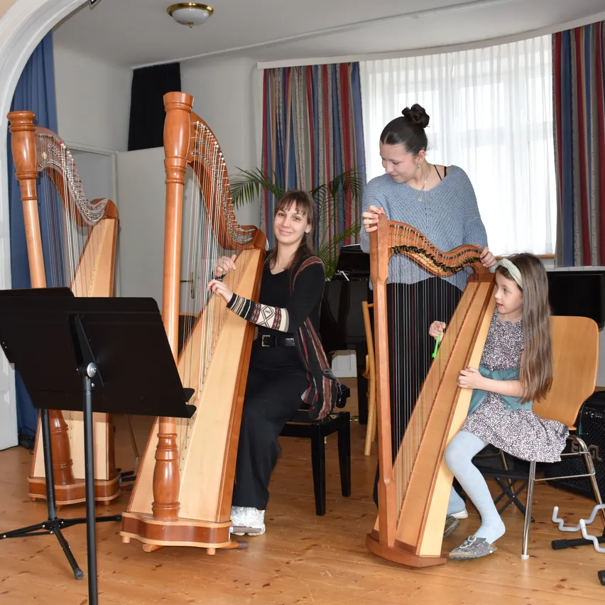 Drei Frauen in einem Raum mit drei Harfen. Eine Frau sitzt und spielt eine Harfe, eine andere steht und spielt eine Harfe, und ein junges Mädchen spielt ebenfalls eine Harfe.