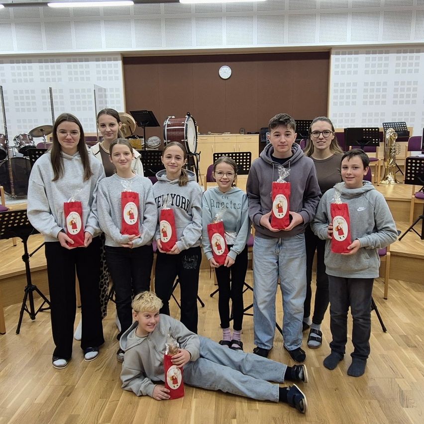 Eine Gruppe von Kindern, die rote Geschenktüten mit einer Weihnachtsfigur halten, stehen und sitzen in einem Musikraum mit Musikinstrumenten im Hintergrund.