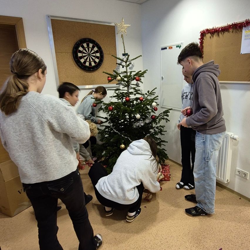 Eine Gruppe von Menschen schmückt einen Weihnachtsbaum in einem Raum mit einem Dartboard und Anschlagtafeln. Zwei Jungen stehen daneben und beobachten, während andere knien, um Geschenke zu arrangieren.