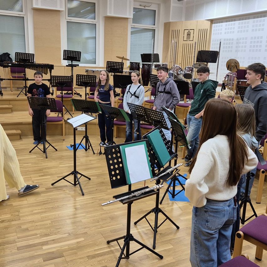 Eine Gruppe von Schülern in einem Musikraum, jeder hält ein Instrument, mit Notenständern und Stühlen im Hintergrund.