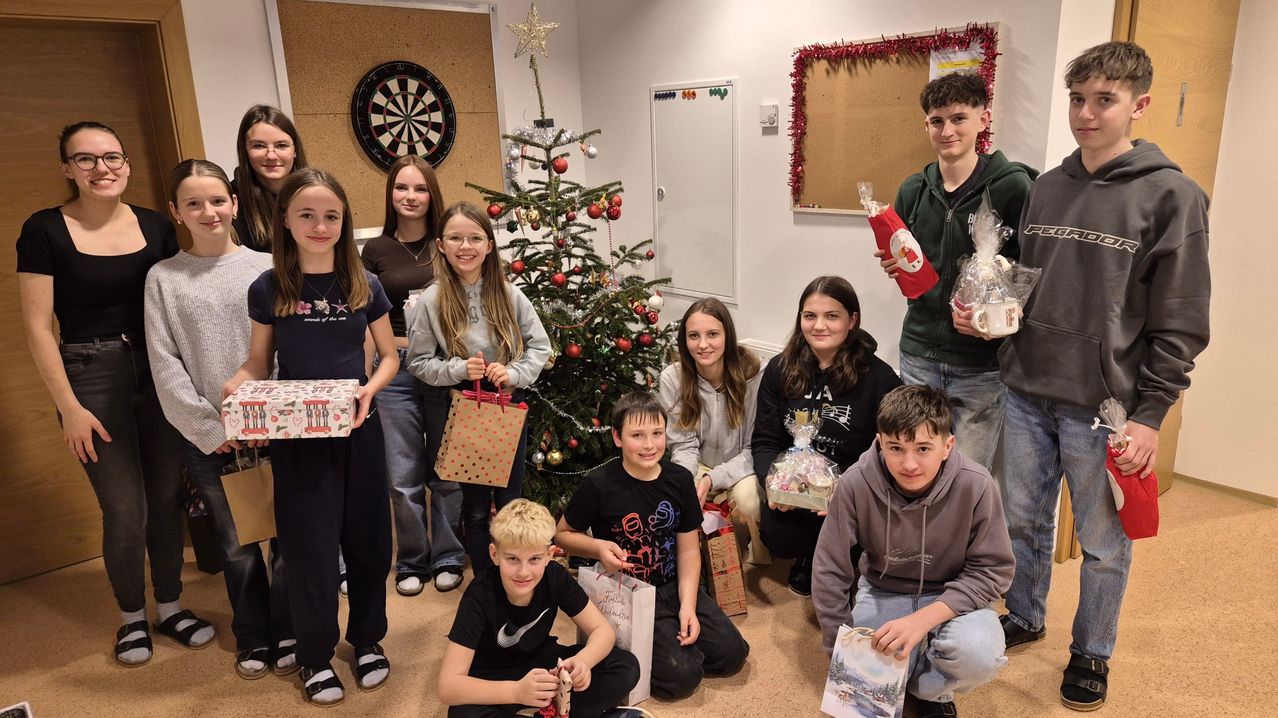 Eine Gruppe von Kindern und Jugendlichen steht um einen geschmückten Weihnachtsbaum herum, lächelt und posiert für ein Foto. Sie halten verschiedene Geschenktüten und verpackte Geschenke. Der Raum ist mit festlichen Dekorationen wie einem Dartboard und einer Pinnwand mit Weihnachtsdekorationen geschmückt.