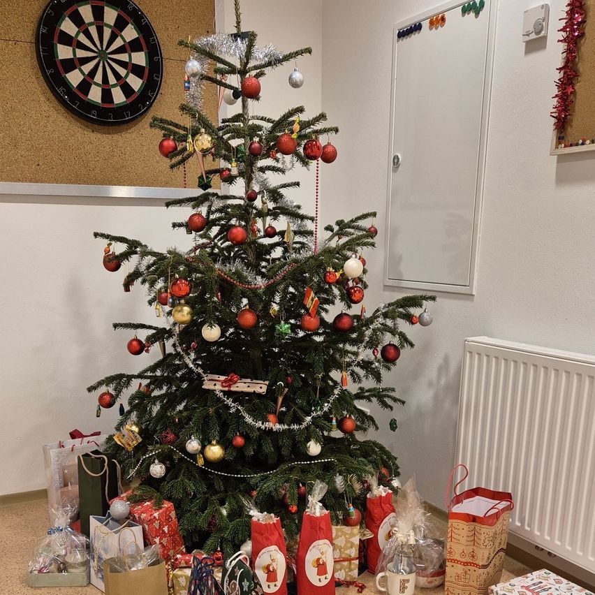 Ein geschmückter Weihnachtsbaum steht in einem Raum mit Geschenken. Dahinter befindet sich ein Dartboard an einer Korkwand und eine weiße Tafel. Rechts steht ein weißer Heizkörper.