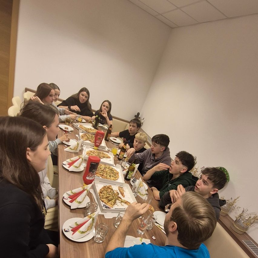Eine Gruppe von Menschen sitzt um einen langen Tisch in einem Raum und genießt Pizza und Getränke. Einige von ihnen schauen in die Kamera, während andere essen. Auf dem Tisch liegen Teller, Flaschen und Servietten.