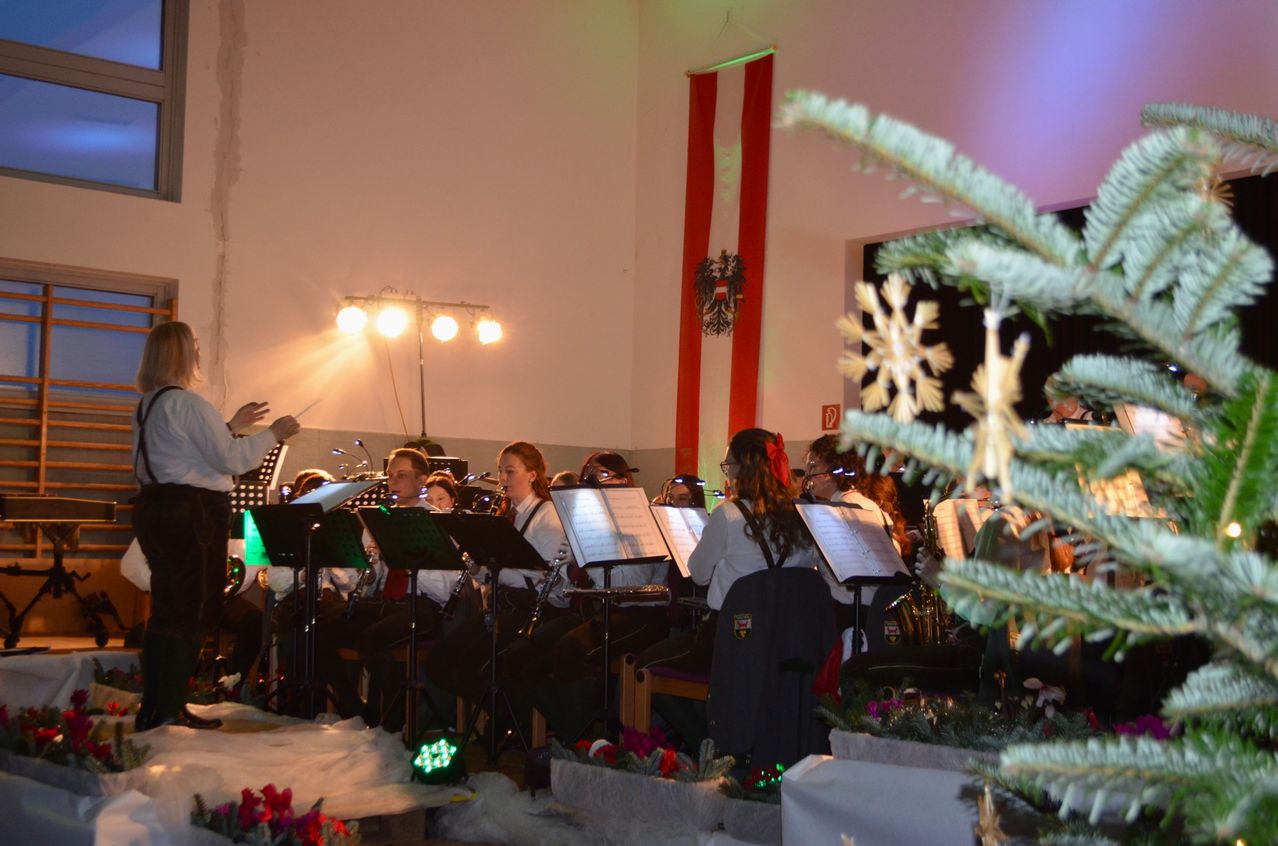 Ein Orchester spielt in einem geschmückten Raum mit roten Flaggen, einem Wappen und Weihnachtsdekorationen. Der Dirigent führt, während die Musiker auf Stühlen mit Notenständern sitzen.