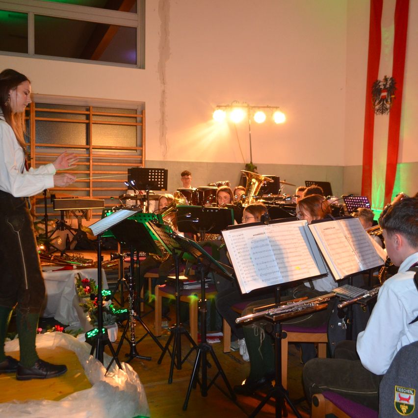 Eine Frau dirigiert eine Band in Innenräumen. Sie spielen Trompeten, Posaunen und andere Instrumente. Das Publikum sitzt auf Stühlen. Der Raum hat eine weiße Wand, einen roten Banner und eine Lampe.