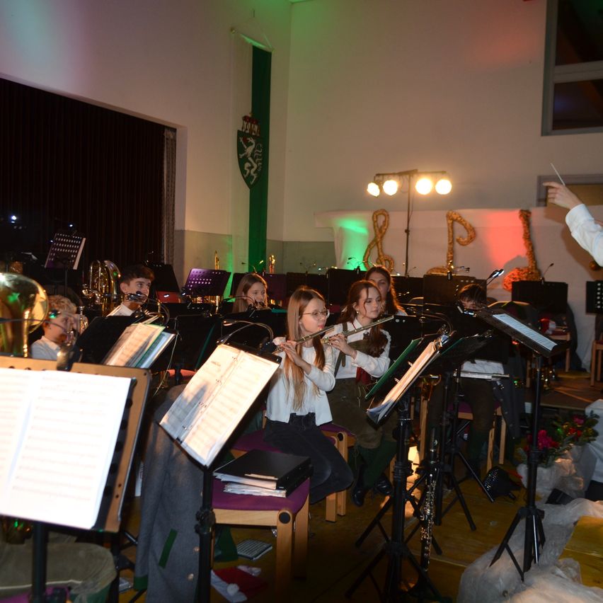 Ein Schulorchester spielt mit einem Dirigenten, der die Schüler anführt. Die Schüler sitzen auf Stühlen und halten Instrumente wie Trompeten, Flöten und Posaunen. Die Bühne ist mit musikalischen Noten und festlichen Lichtern geschmückt.