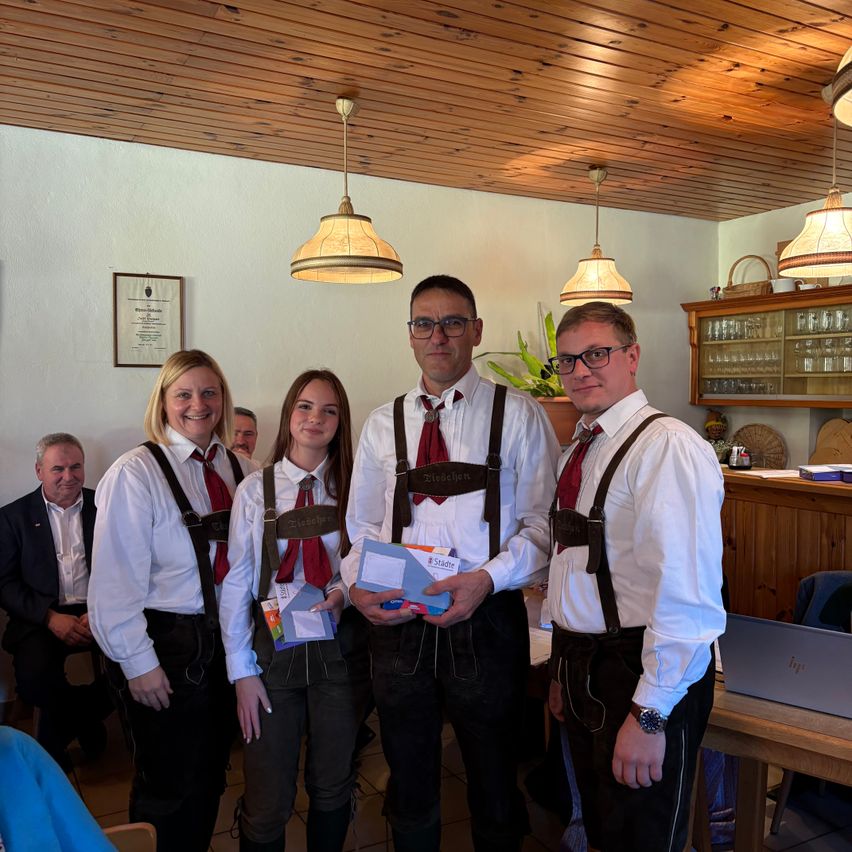 Fünf Personen in traditioneller deutscher Kleidung stehen in einem Raum und halten verschiedene Gegenstände. Zwei Männer und zwei Frauen tragen Hosenträger und rote Krawatten. Der Mann auf der rechten Seite hält eine blaue Mappe und eine Schachtel. Hinter ihnen sitzen ein weiterer Mann in einem Anzug und eine Frau in einem Kleid auf einem Stuhl.