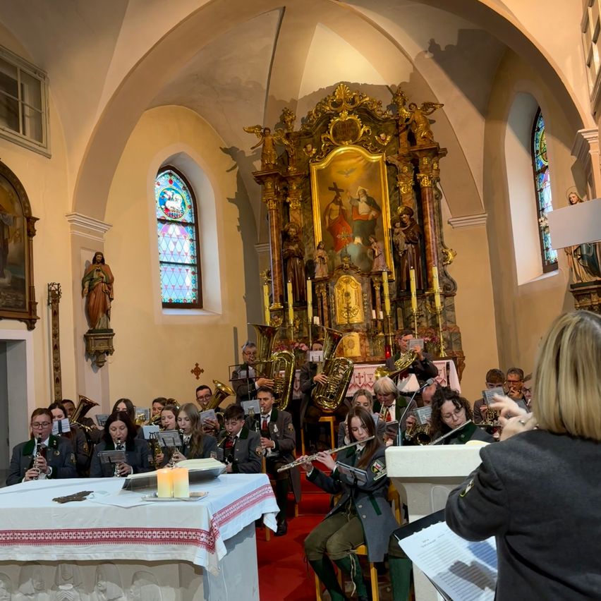 Ein Bläserensemble spielt in einer Kirche. Die Musiker sitzen vor einem Tisch mit Kerzen, und der Dirigent steht rechts. Dahinter befindet sich ein Altar mit goldenem Rahmen und einer Statue einer Heiligen.
