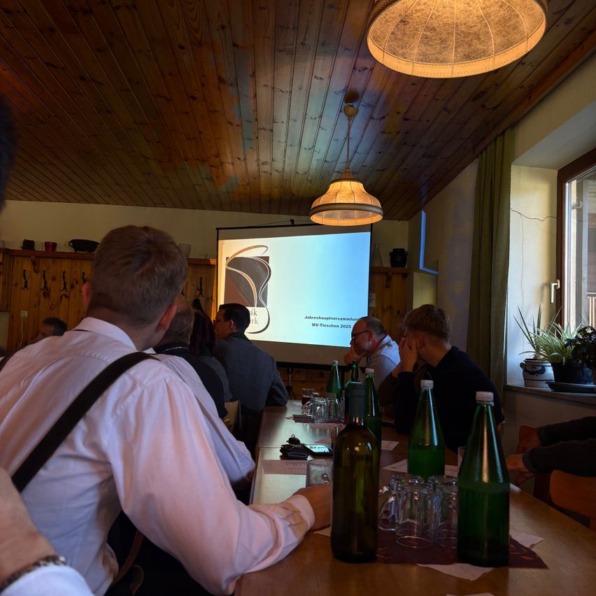 Eine Gruppe von Menschen sitzt um einen Tisch in einem Raum mit Holzwänden, einer Projektionsleinwand und einer Lampe. Einige schauen sich eine Präsentation an, während andere ihre Telefone betrachten. Flaschen und Gläser stehen auf dem Tisch.