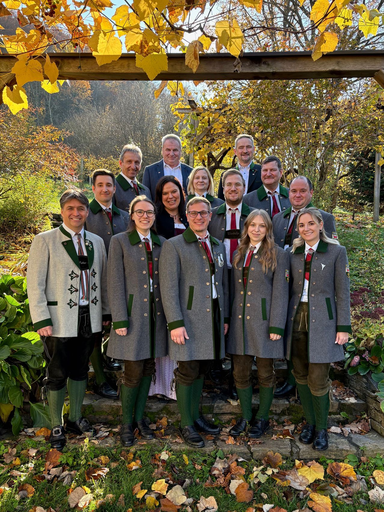 Eine Gruppe von Menschen in traditioneller bayerischer Kleidung, einschließlich Lederhosen und Jacken mit grünem Besatz, steht in einem Garten mit Bäumen und Pflanzen im Hintergrund. Sie posieren für ein Foto.