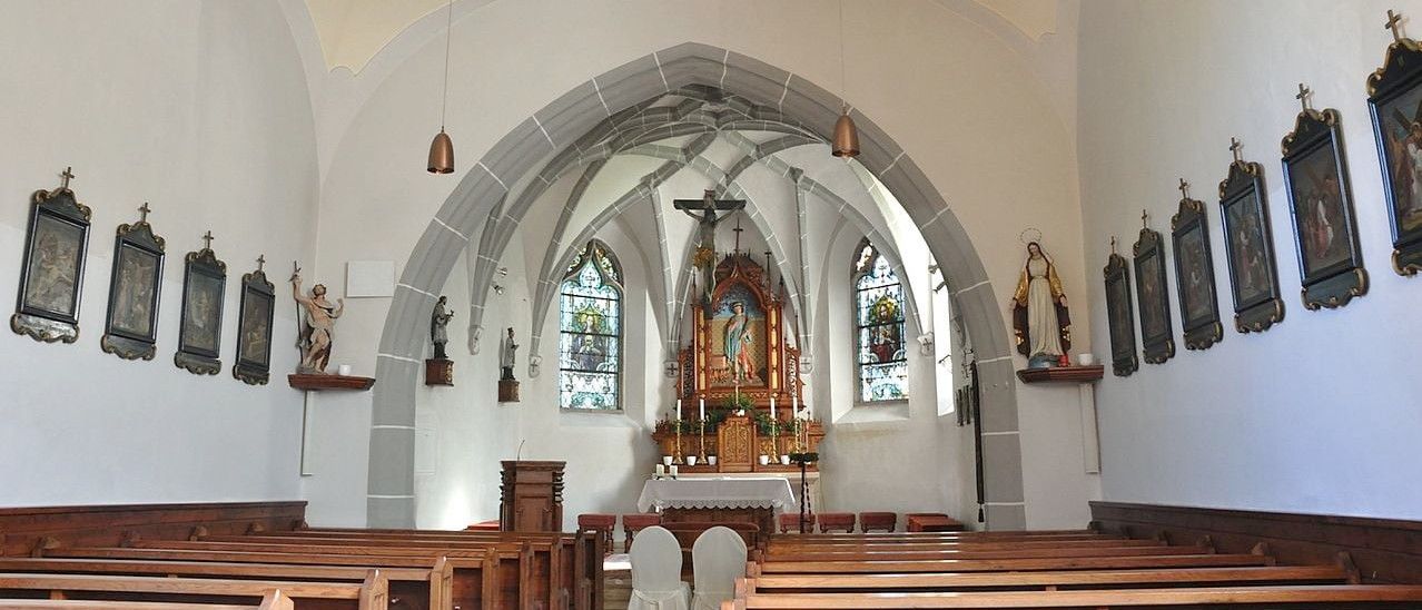 Ein Innenraum einer Kirche mit gotischem Bogen, Altar und Statuen. Holzbänke säumen beide Seiten.
