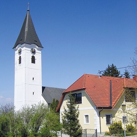 Eine Kirche mit einem hohen weißen Turm mit einem Spitzturm und zwei Uhren befindet sich neben einem gelben Haus mit einem roten Dach.