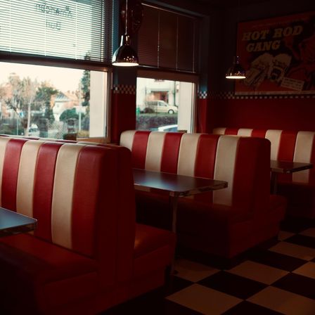 Ein leerer Diner mit rot-weiß karierten Sitzbänken, einem karierten Boden und großen Fenstern, die einen Blick auf geparkte Autos und Bäume zeigen.