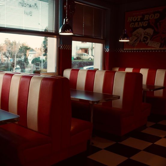 Ein leerer Diner mit rot-weiß karierten Sitzbänken, einem karierten Boden und großen Fenstern, die einen Blick auf geparkte Autos und Bäume zeigen.