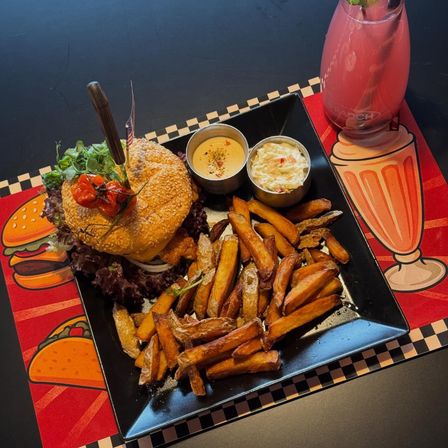 Ein Burger mit einem Messer, Salat und einer Tomate sitzt auf einem schwarzen quadratischen Teller. Er wird von einer Saucenschüssel und Pommes Frites begleitet. Ein Glas mit einer rosa Flüssigkeit und einem Strohhalm steht rechts.