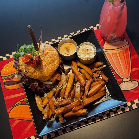 Ein Burger mit einem Messer, Salat und einer Tomate sitzt auf einem schwarzen quadratischen Teller. Er wird von einer Saucenschüssel und Pommes Frites begleitet. Ein Glas mit einer rosa Flüssigkeit und einem Strohhalm steht rechts.