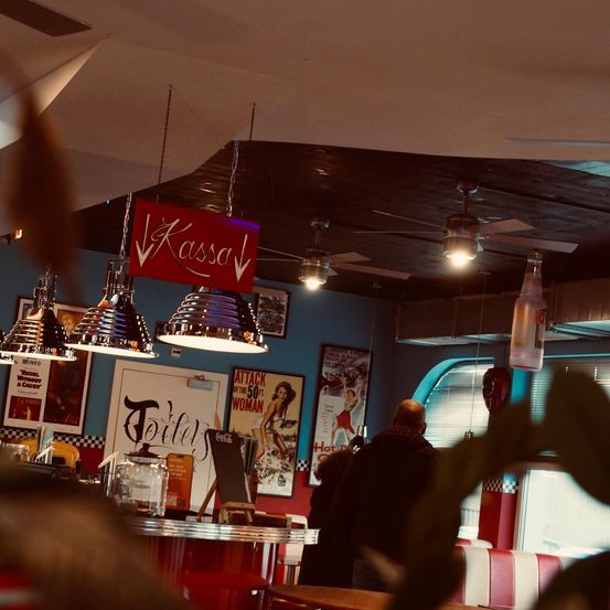 Ein Indoor-Imbiss mit einem roten Schild, das Kassa über der Theke sagt, Menschen auf einer Bank sitzend und Poster an der Wand.