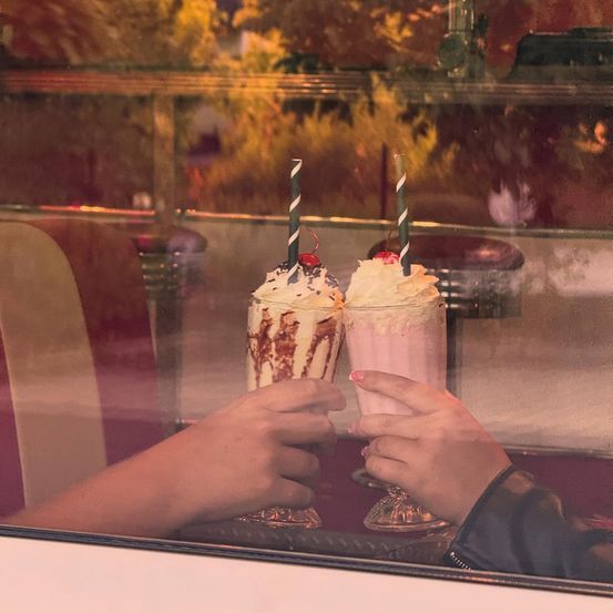 Zwei Hände stoßen mit Milchshakes in einem Retro-Diner an, mit einer Kirsche und Streuseln obenauf. Ein Spiegelbild von Bäumen und Stühlen ist im Glas zu sehen.