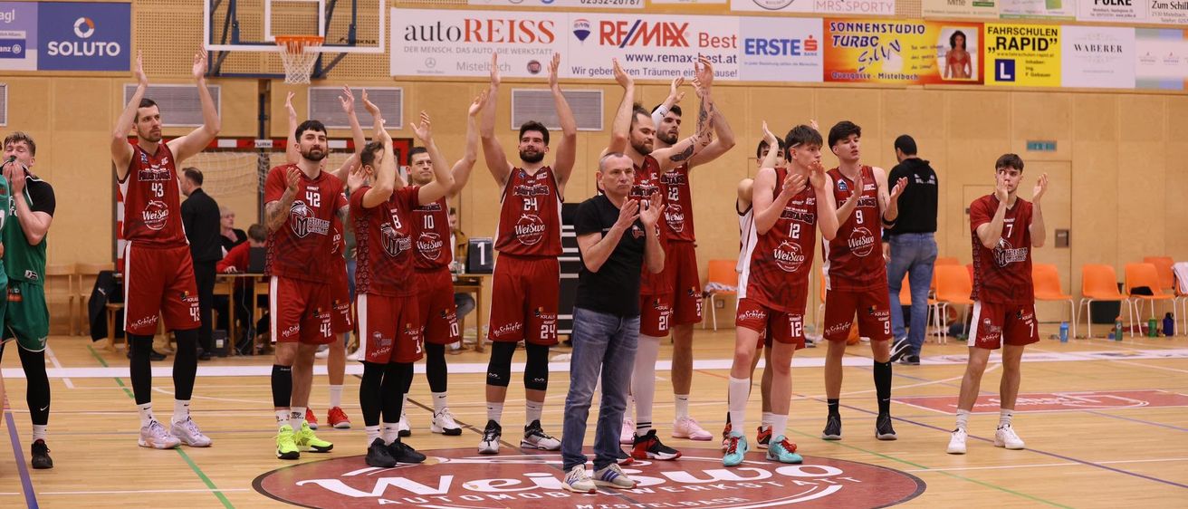 Ein Basketballteam feiert auf einem Spielfeld mit dem Trainer davor, umgeben von einem jubelnden Publikum. Verschiedene Werbebanner schmücken die Wände.