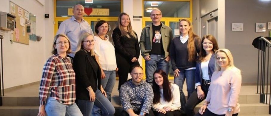 Eine Gruppe von Menschen, sowohl Männer als auch Frauen, posiert für ein Foto auf den Stufen. Sie tragen lässige Kleidung und lächeln. Hinter ihnen befinden sich Glastüren mit einem gelben Rand.