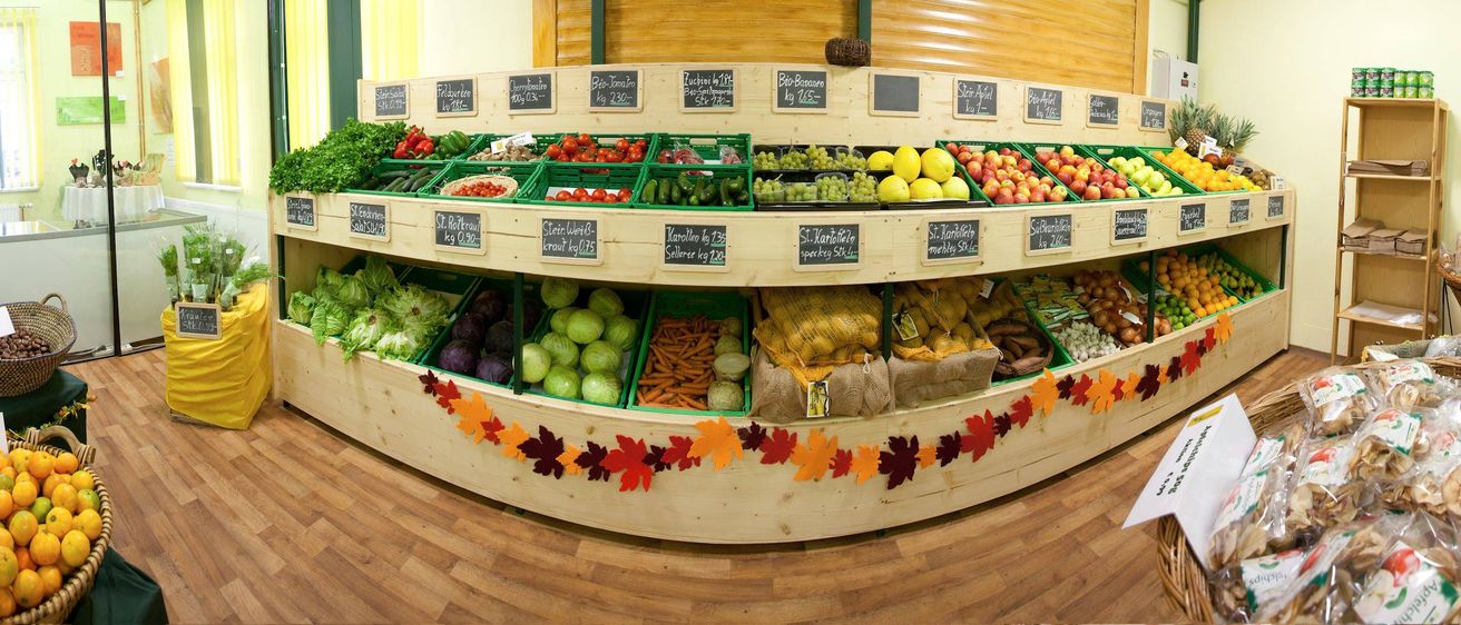 Eine hölzerne Präsentation zeigt verschiedene Gemüsesorten wie Gurken, Tomaten und Karotten. Jedes Gemüse ist mit seinem Namen und Preis beschriftet. Die Präsentation ist mit Herbstblättern dekoriert.