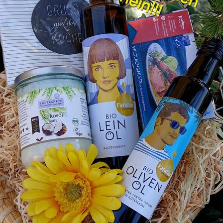Ein Korb enthält eine Flasche Bio-Olivenöl, eine Flasche Leinöl, ein Glas Kokosöl und eine gelbe Blume. Ein roter und weißer Karton mit der Aufschrift 'Wildlachsfilets' befindet sich neben dem Korb. Ein runder Aufkleber mit der Aufschrift 'Gruss aus der Kuche' befindet sich links.