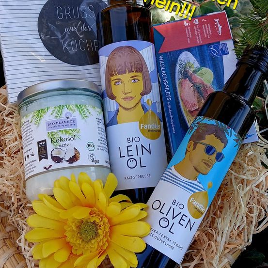 Ein Korb enthält eine Flasche Bio-Olivenöl, eine Flasche Leinöl, ein Glas Kokosöl und eine gelbe Blume. Ein roter und weißer Karton mit der Aufschrift 'Wildlachsfilets' befindet sich neben dem Korb. Ein runder Aufkleber mit der Aufschrift 'Gruss aus der Kuche' befindet sich links.
