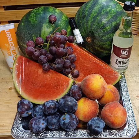 Ein silbernes Tablett zeigt Früchte: Wassermelone, Trauben, Pflaumen und Pfirsiche. Eine Flasche Knobl Schnaps und Sonnencreme sind ebenfalls auf dem Tisch.
