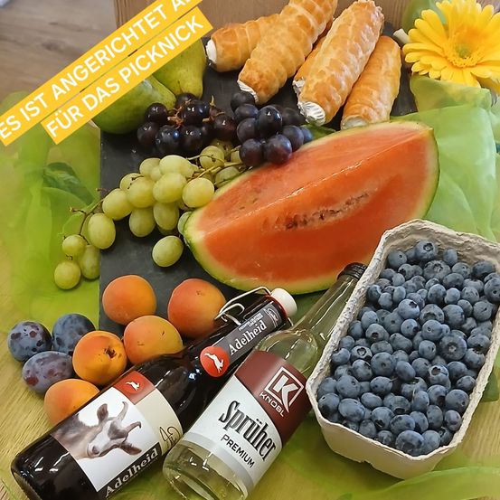 Auf einem Tisch befinden sich Obst wie Äpfel, Birnen, Trauben und Blaubeeren. Es gibt auch zwei Flaschen und eine geschnittene Wassermelone. Der Text lautet 'Wut ist als Snack serviert.'