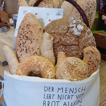 Ein Korb mit verschiedenen Brotsorten, einschließlich Vollkorn-, Sesam- und Bagelbrot, auf einem Holztisch. Vorne steht ein Beutel mit dem Zitat 'Der Mensch lebt nicht vom Brot allein'.