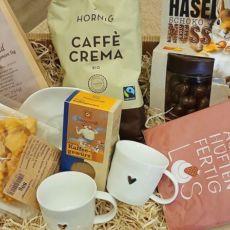 Eine Box enthält verschiedene Artikel, darunter eine weiße Tasse, eine weiße Schüssel, ein Glas und ein Buch. Andere Artikel sind eine Kaffeesahne, ein Beutel Kaffeebohnen, ein Beutel Haselnüsse und eine kleine Packung Kekse.