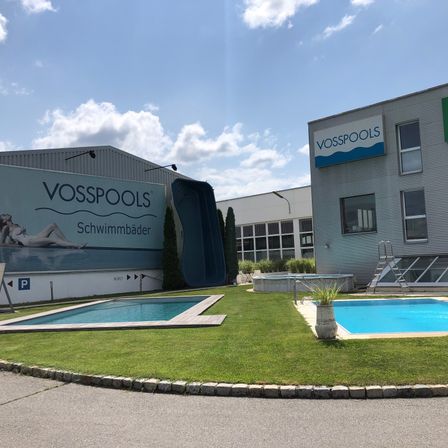 Ein Vosspools-Showroom zeigt zwei Schwimmbecken, einen Rasen und ein Gebäude mit einem Schild und einer Rutsche. Ein Becken hat eine Topfpflanze.