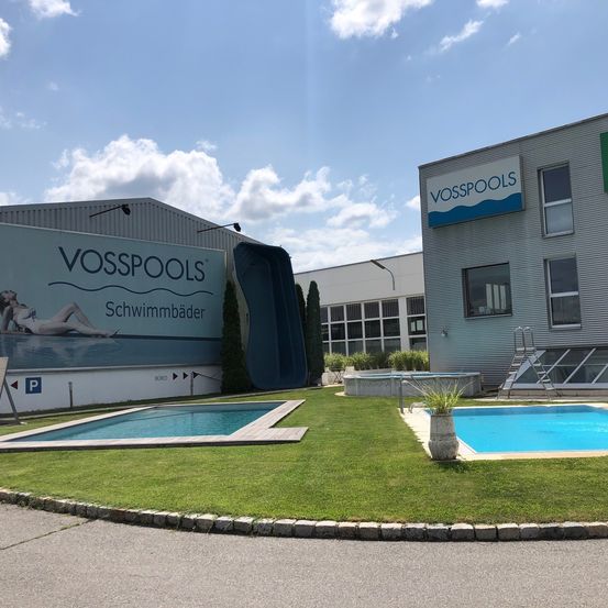 Ein Vosspools-Showroom zeigt zwei Schwimmbecken, einen Rasen und ein Gebäude mit einem Schild und einer Rutsche. Ein Becken hat eine Topfpflanze.