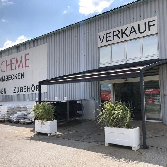 Die Vorderseite eines Verkaufsgebäudes mit einem Vordach über dem Eingang, zwei Topfpflanzen und einem Schild, das Verkauf sagt.