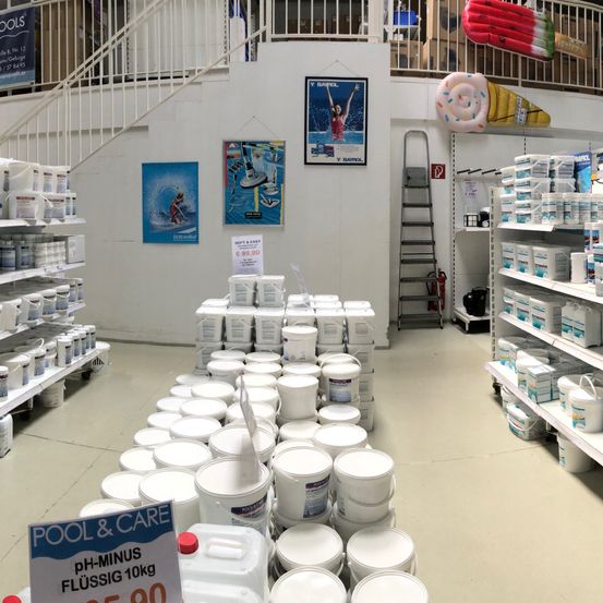Ein Poolbedarfsladen mit weißen Regalen, Eimern und Kanistern. Eine Leiter, ein Feuerlöscher und Poster an der Wand. Eine Treppe führt in den zweiten Stock.