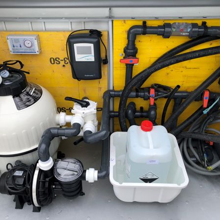 Ein Pump- und Filtersystem für einen Pool ist an einer gelben Wand befestigt. Ein Wasserkrug und verschiedene Werkzeuge sind in der Nähe.