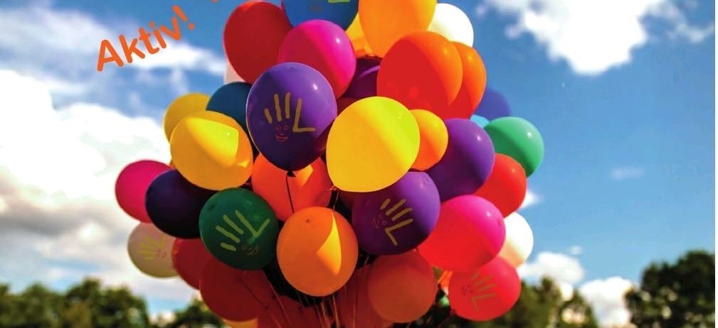Eine lebhafte Parkszene mit einem bunten Ballonbouquet im Vordergrund, einem bewölkten Himmel und einem Rasenfeld. Die Worte 'Aktiv! Kreativ! Schulfest!' sind oben zu sehen.