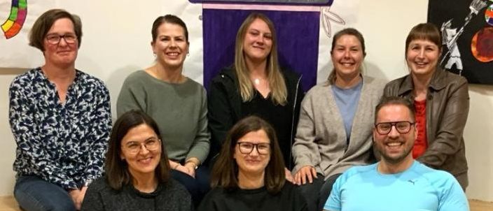 Eine Gruppe von acht Menschen, einschließlich Männer und Frauen, sitzen auf einer Holzbank. Sie alle lächeln für ein Foto. Hinter ihnen hängen bunte Banner.