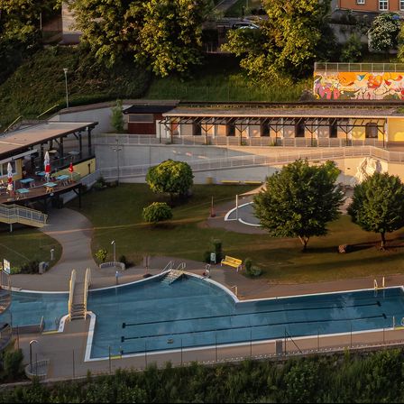 Eine Luftaufnahme eines Schwimmbeckens mit einem farbenfrohen Wandbild an der Seite. Bäume, eine gelbe Bank und Regenschirme sind sichtbar. Gebäude und ein Weg sind im Hintergrund.