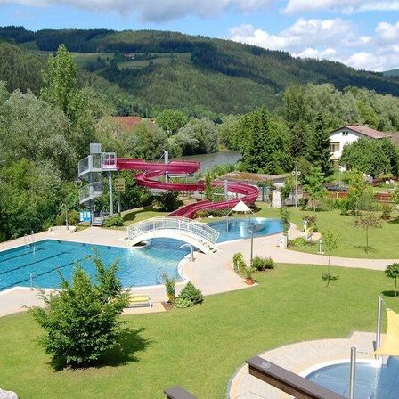 Ein Luftbild eines Wasserparks mit einer großen roten Wasserrutsche, einer Brücke und einem Schwimmbecken. Bäume und Pflanzen umgeben das Gebiet. Im Hintergrund befinden sich Häuser und ein Fluss.
