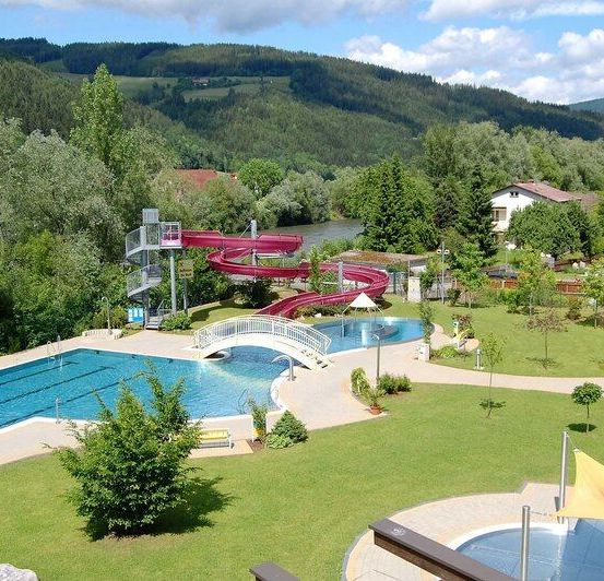 Ein Luftbild eines Wasserparks mit einer großen roten Wasserrutsche, einer Brücke und einem Schwimmbecken. Bäume und Pflanzen umgeben das Gebiet. Im Hintergrund befinden sich Häuser und ein Fluss.